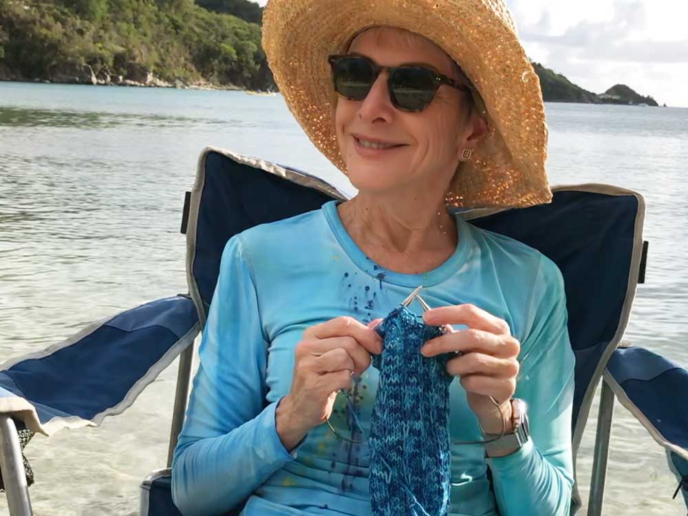 woman in a hat and sunglasses sitting in a beach chair  on the beach while she knits